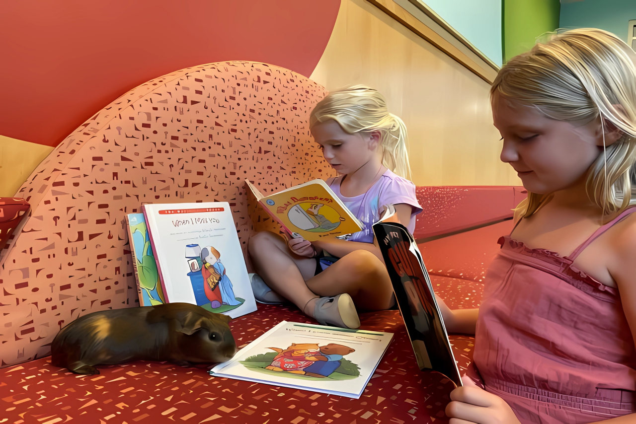 Two young children holding picture books reading to a brown guinea pigs while seated on some cushions